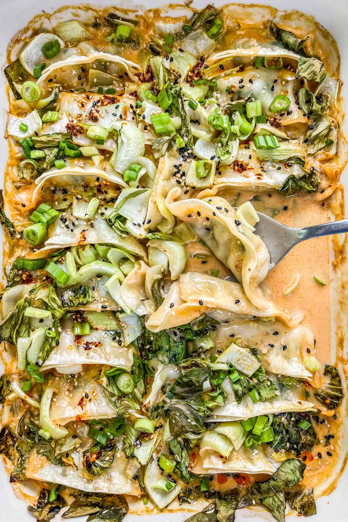 Casserole dish with baked dumplings and coconut milk topped with green onions, bok choy, sesame seeds, and chili oil and a silver spoon scooping them up.