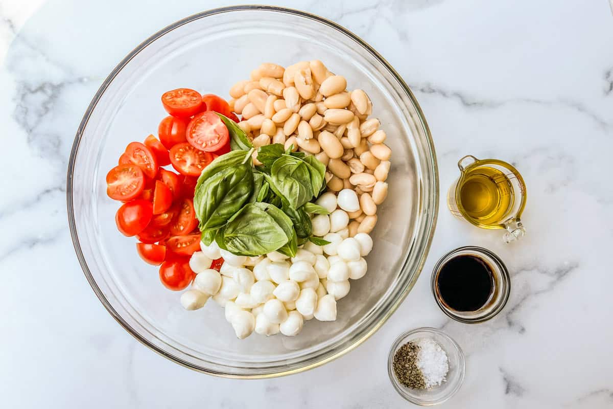 White beans, cherry tomatoes, mozzarella balls, and basil leaves in a large glass bowl next to bowls of olive oil, balsamic, and salt and pepper.