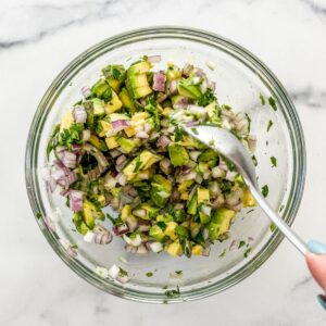 An avocado red onion salsa being mixed in a small glass bowl with a spoon.