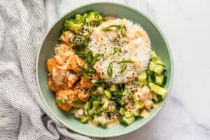 A bang bang salmon bowl with rice, cucumbers, avocado, and edamame in a green bowl.