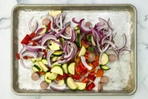 Sliced sausage, zucchini, red onion, and bell peppers in a pile on a silver sheet pan.