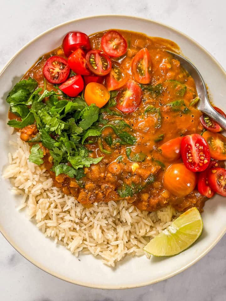 Coconut lentil curry with rice, topped with cilantro and sliced tomatoes, in a white bowl with a silver spoon.