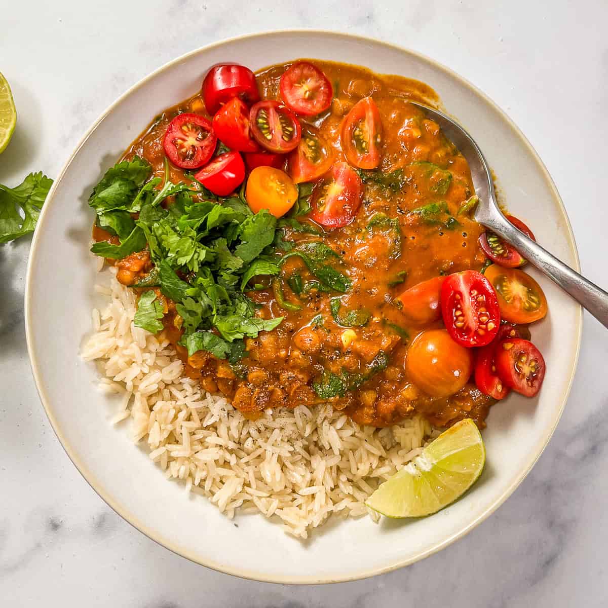 Coconut lentil curry with rice, topped with cilantro and sliced tomatoes, in a white bowl with a silver spoon.