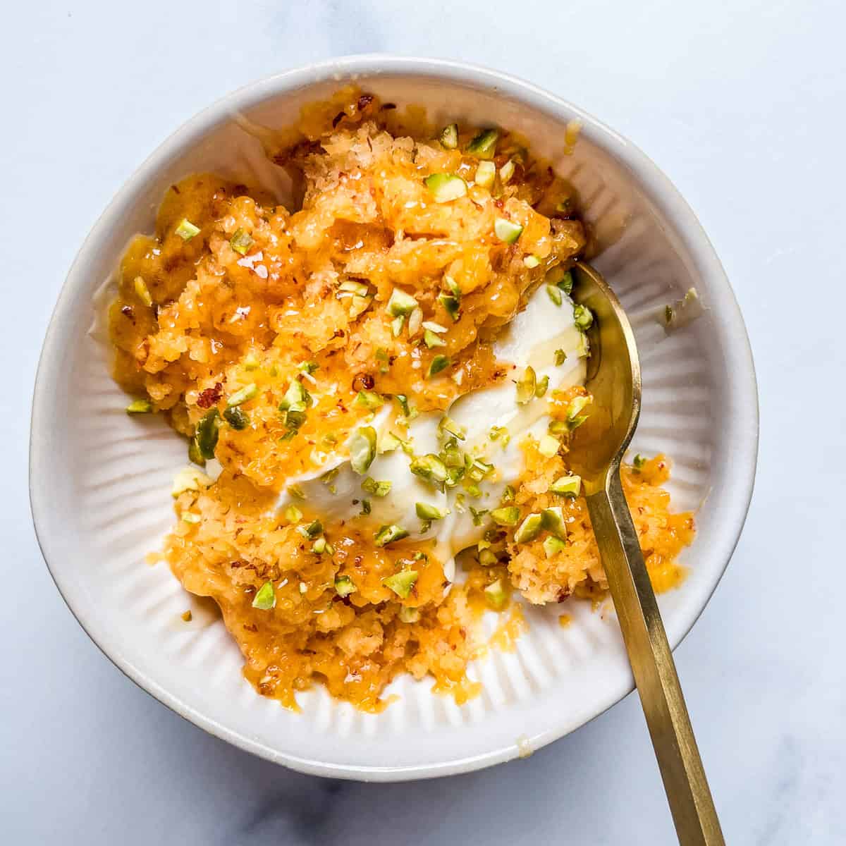 Shaved frozen peach with mascarpone, honey, and pistachios in a white bowl with a gold spoon.