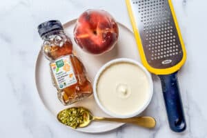 Honey, a frozen peach, mascarpone, a spoon holding chopped pistachios, and a cheese grater on a white plate.