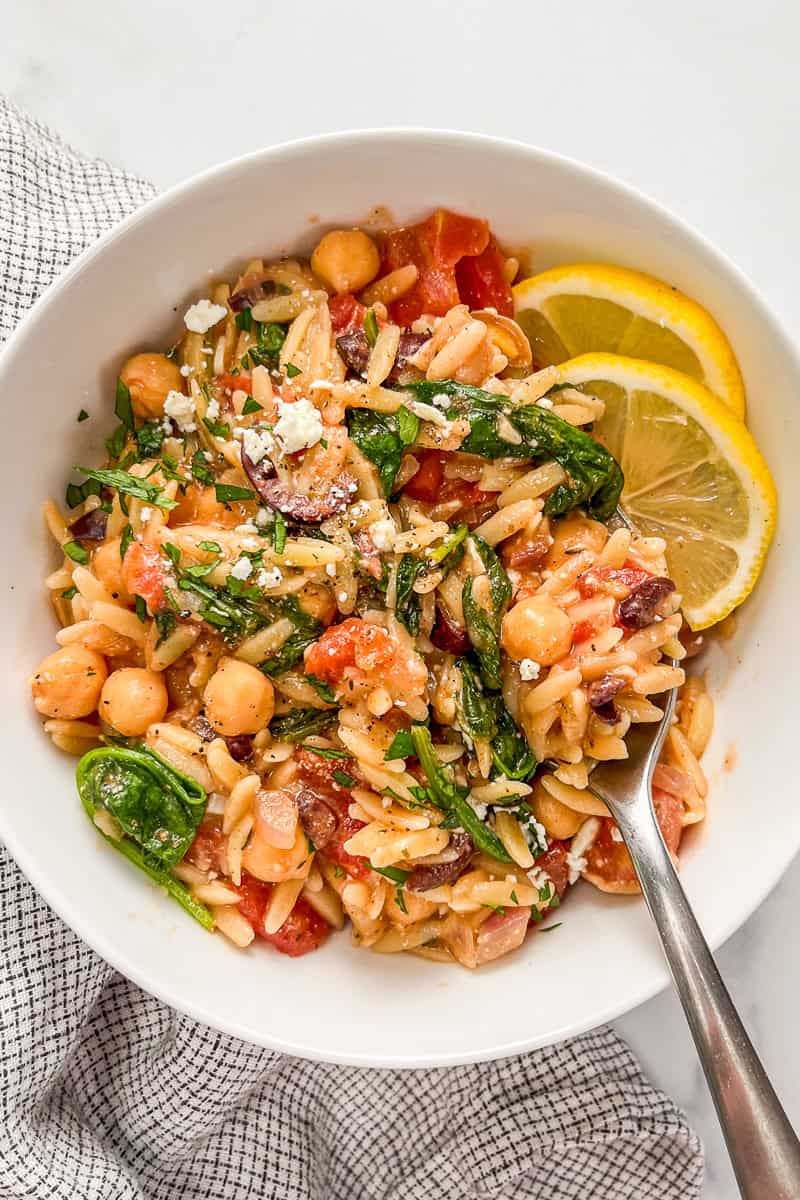 Mediterranean chickpea orzo bake with spinach in a white bowl with lemon slices and a silver spoon.