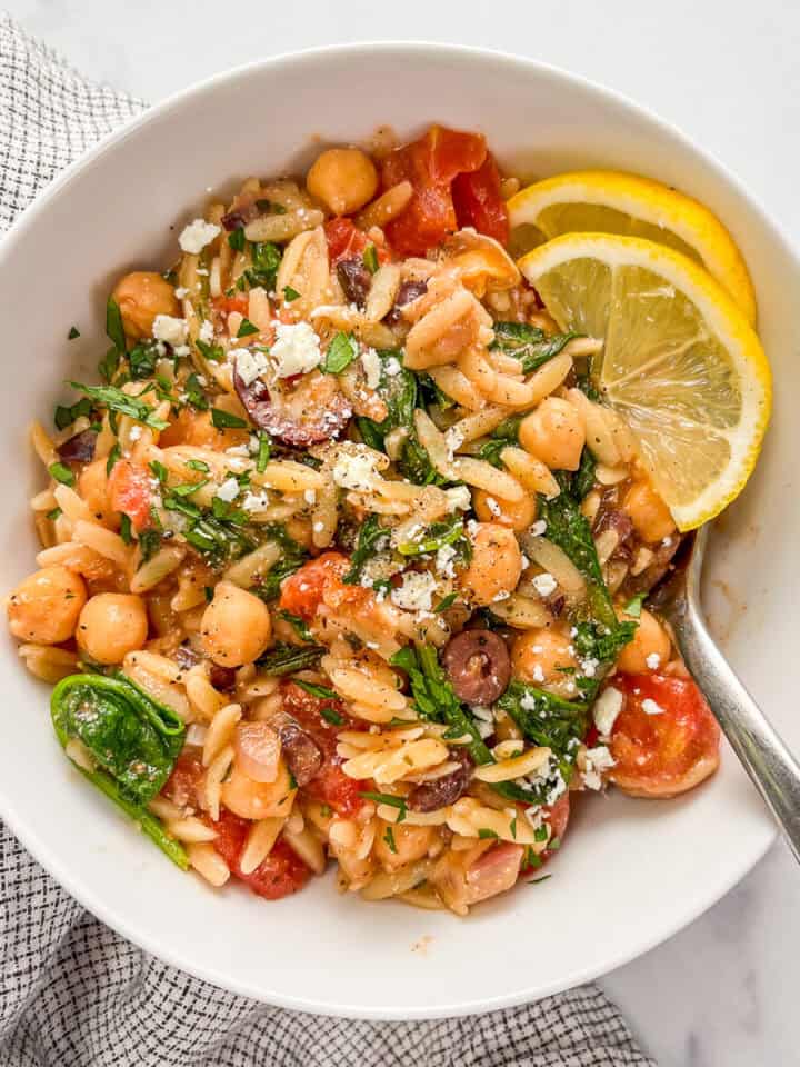 Mediterranean chickpea orzo bake with spinach in a white bowl with lemon slices and a silver spoon.