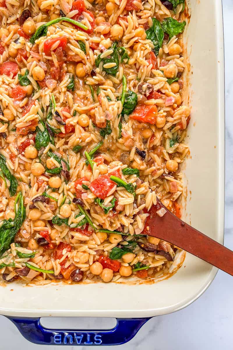 Mediterranean chickpea orzo bake with spinach and tomatoes in a baking dish wit a wooden spoon.