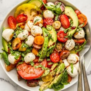 An avocado caprese salad in a white serving bowl with a fork and spoon.