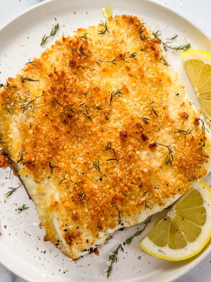Baked parmesan panko halibut on a white plate with slices of lemon.