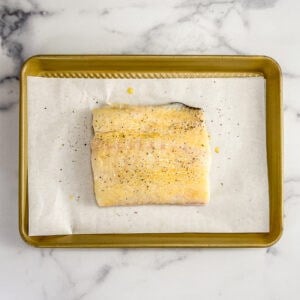 A piece of halibut brushed with butter and sprinkled with salt and pepper on a parchment lined sheet pan.