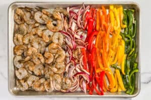 Green, yellow, orange, and red bell peppers, red onions, and shrimp topped with spices on a sheet pan.