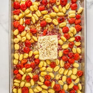 A block of baked feta surrounded by tomatoes, gnocchi, and red onion on a silver sheet pan.