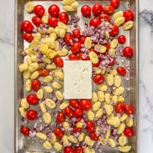 A block of feta surrounded by tomatoes, gnocchi, and red onion on a silver sheet pan.