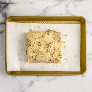A piece of halibut topped with a mixture of panko, parmesan, and thyme on a parchment lined sheet pan.