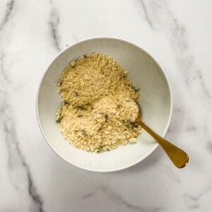 A white bowl of panko, parmesan, and thyme with a gold spoon.