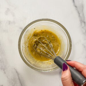 Miso, tahini, olive oil, salt, and Aleppo pepper being mixed in a glass bowl with a whisk.