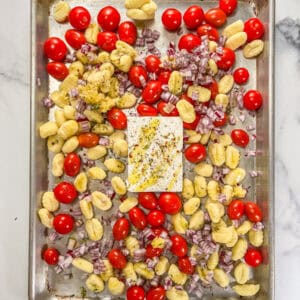 A block of feta topped with oil and spices, surrounded by tomatoes, gnocchi, and red onion on a silver sheet pan.