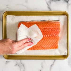 A salmon fillet on a parchment line sheet pan being patted with a paper towel.