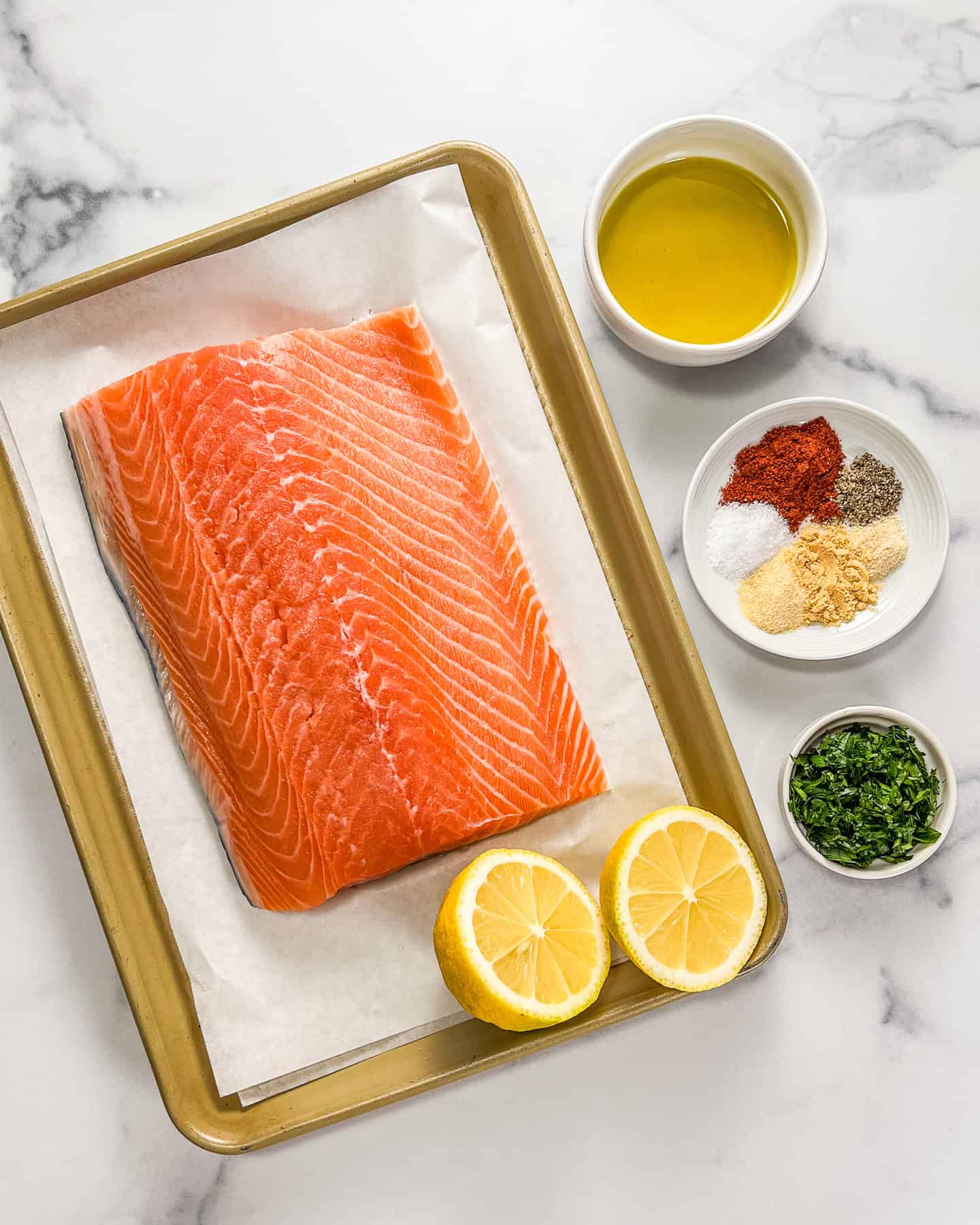 Salmon fillet, a halved lemon, chopped parsley, a small plate of seasonings, and olive oil on a marble background.