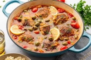 Moroccan chicken with tomatoes, lemon, and green olives in a pot surrounded by herbs, tomatoes, and couscous.