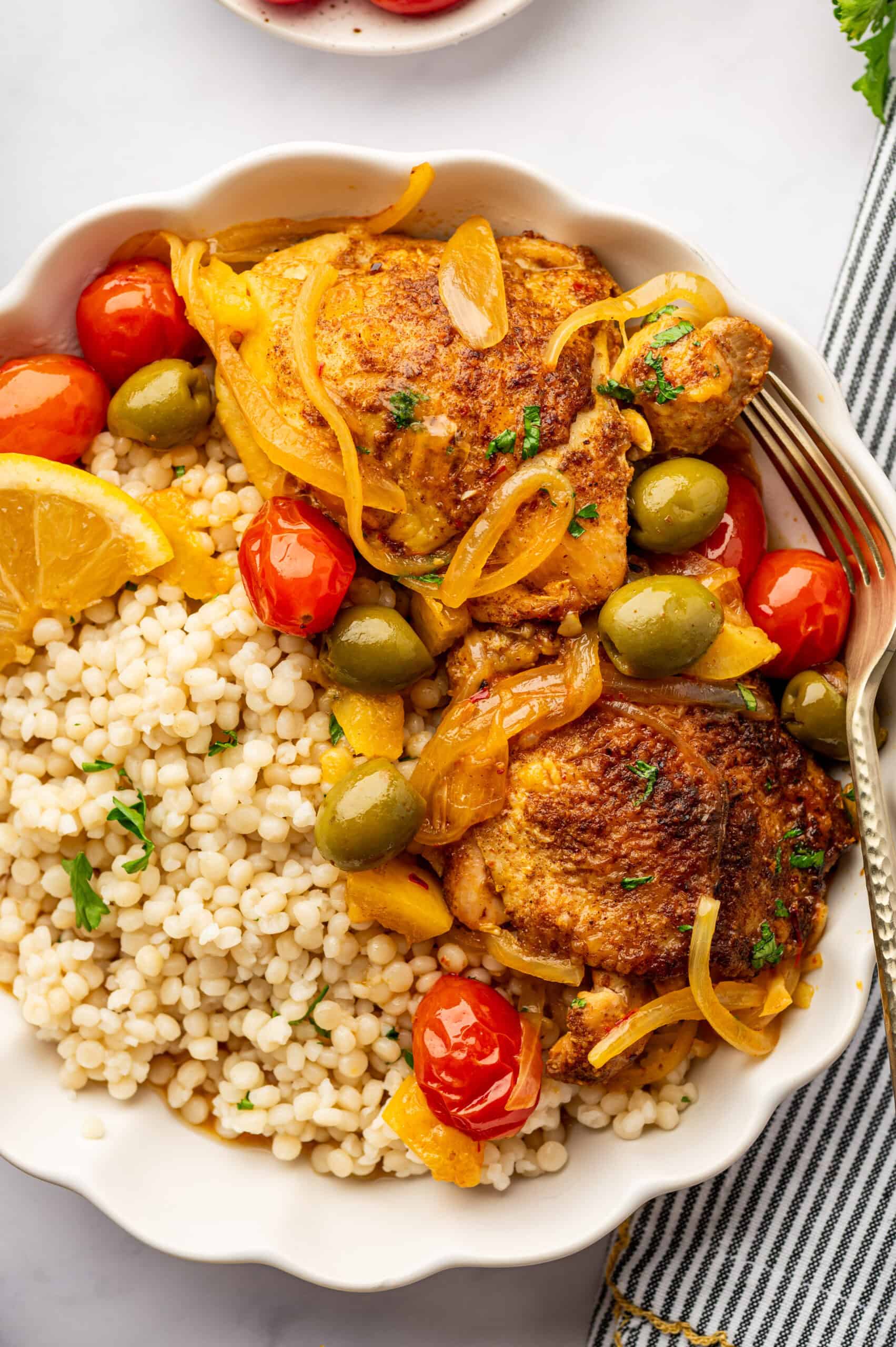 Moroccan chicken with tomatoes, lemon, and green olives in a bowl with couscous with a gold fork.