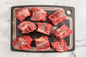 Beef chunks topped with salt and pepper on a black cutting board.