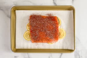 Seasoned salmon fillet on top of lemon slices and a piece of parchment paper on a gold sheet pan.
