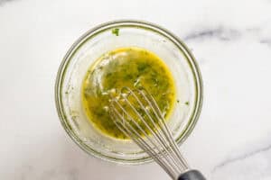 A glass bowl with melted butter and herbs being stirred with a whisk.