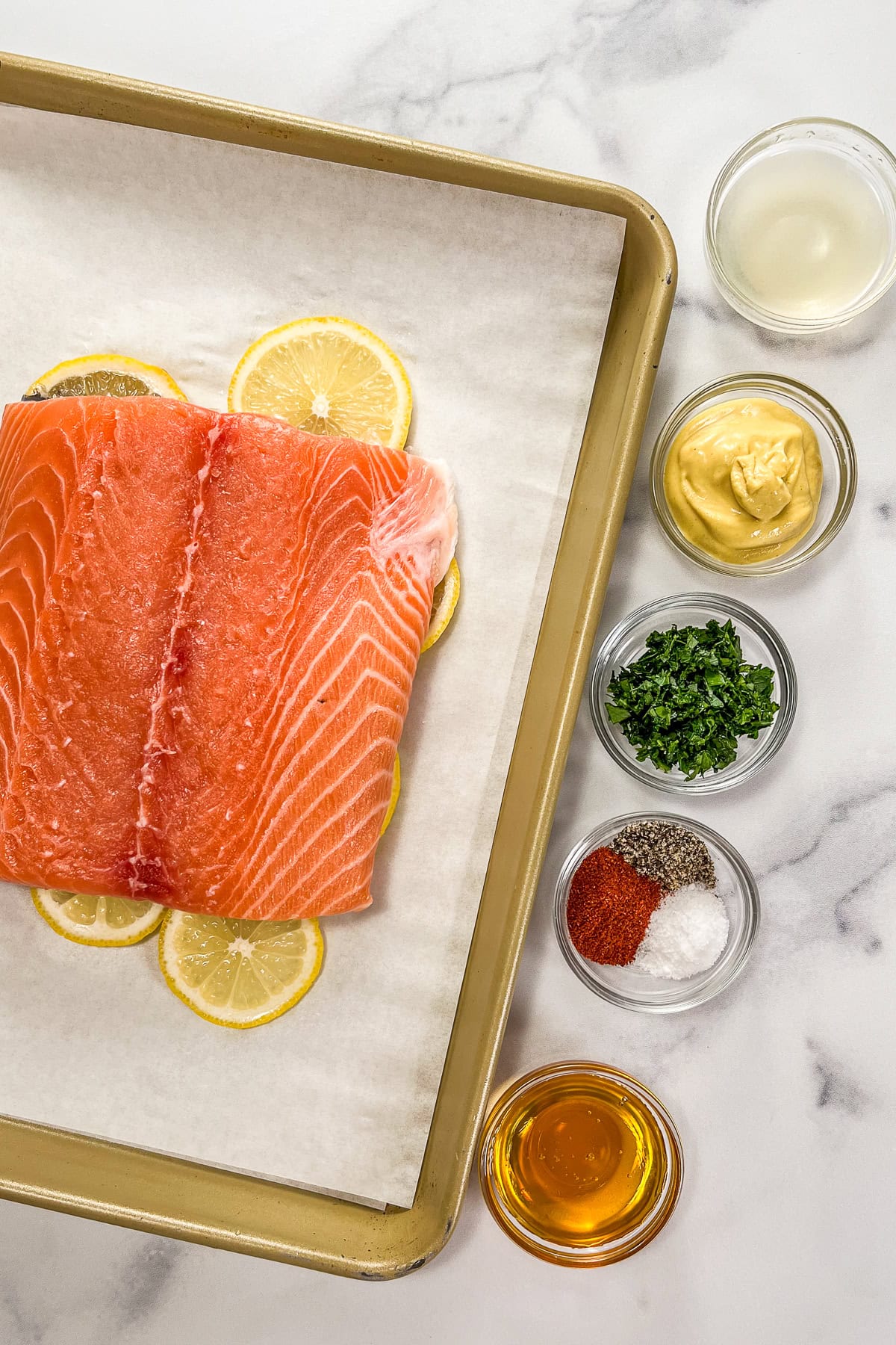 A salmon fillet on top of lemon slices and a piece of parchment paper on a sheet pan, next to glas bowls with lemon juice, Dijon mustard, chopped parsley, spices, and honey.