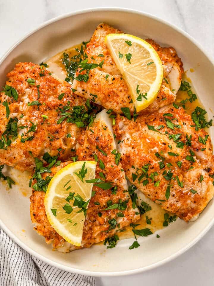 Lemon garlic butter baked cod fillets in a white bowl, topped with lemon slices and parsley.
