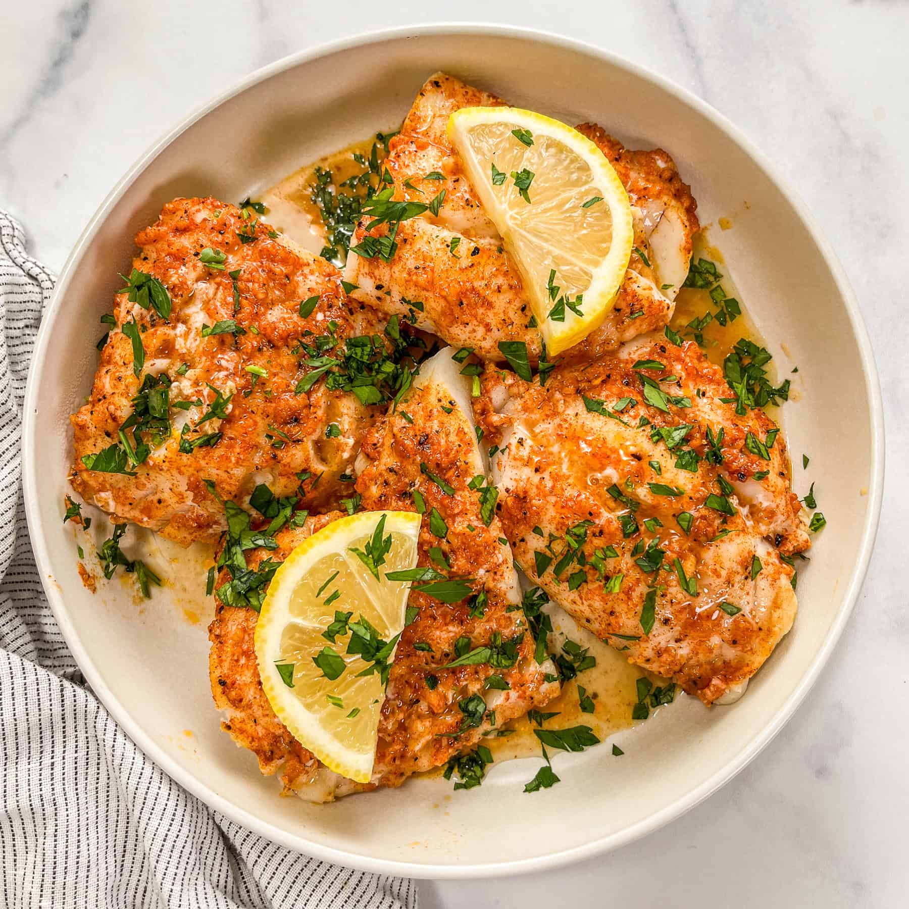 Lemon garlic butter baked cod fillets in a white bowl, topped with lemon slices and parsley.