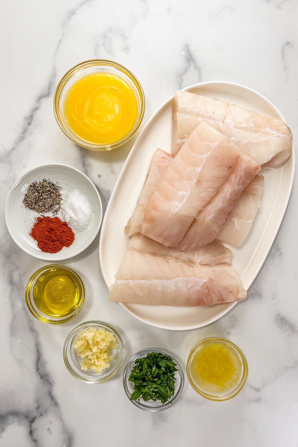 Melted butter, spices, olive oil, minced garlic, chopped parsley, lemon juice and zest, and cod fillets on a marble background.