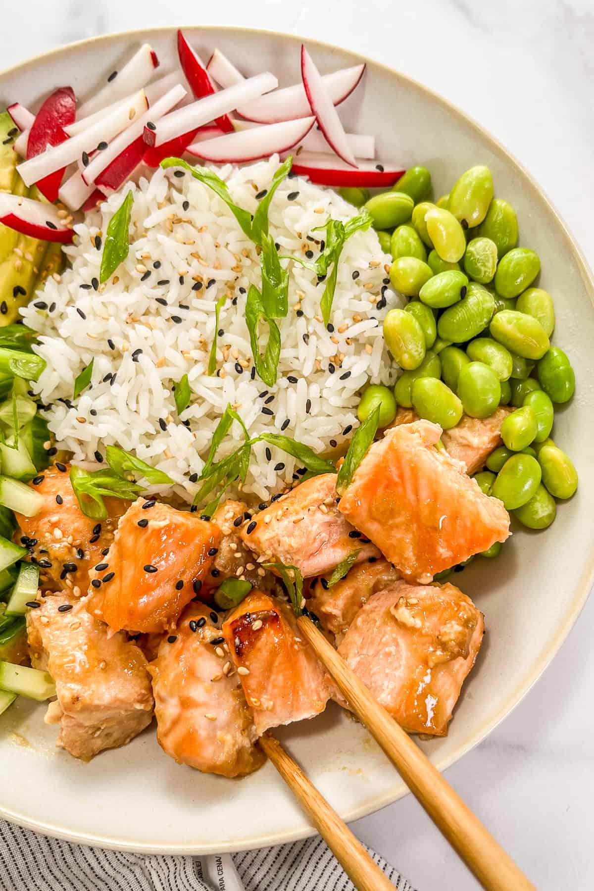 Miso salmon rice bowl with edamame, sliced green onions, sliced radishes, and a pair of wooden chopsticks.