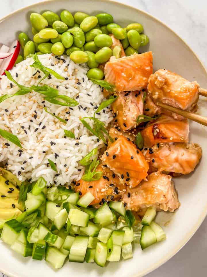 Miso salmon rice bowl with edamame, sliced green onions, sliced radishes, sliced avocado, chopped cucumber, and a pair of wooden chopsticks.