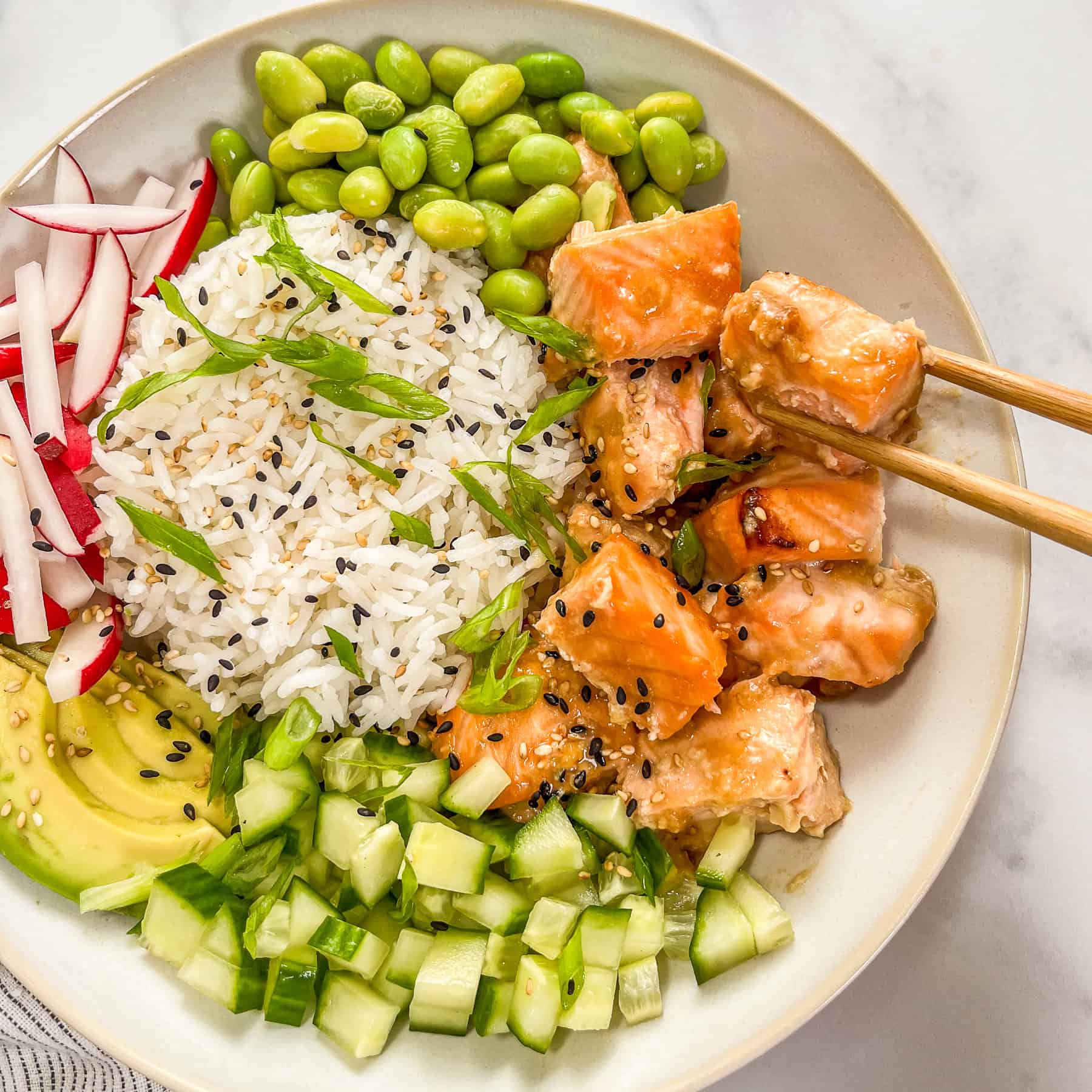 Miso salmon rice bowl with edamame, sliced green onions, sliced radishes, sliced avocado, chopped cucumber, and a pair of wooden chopsticks.