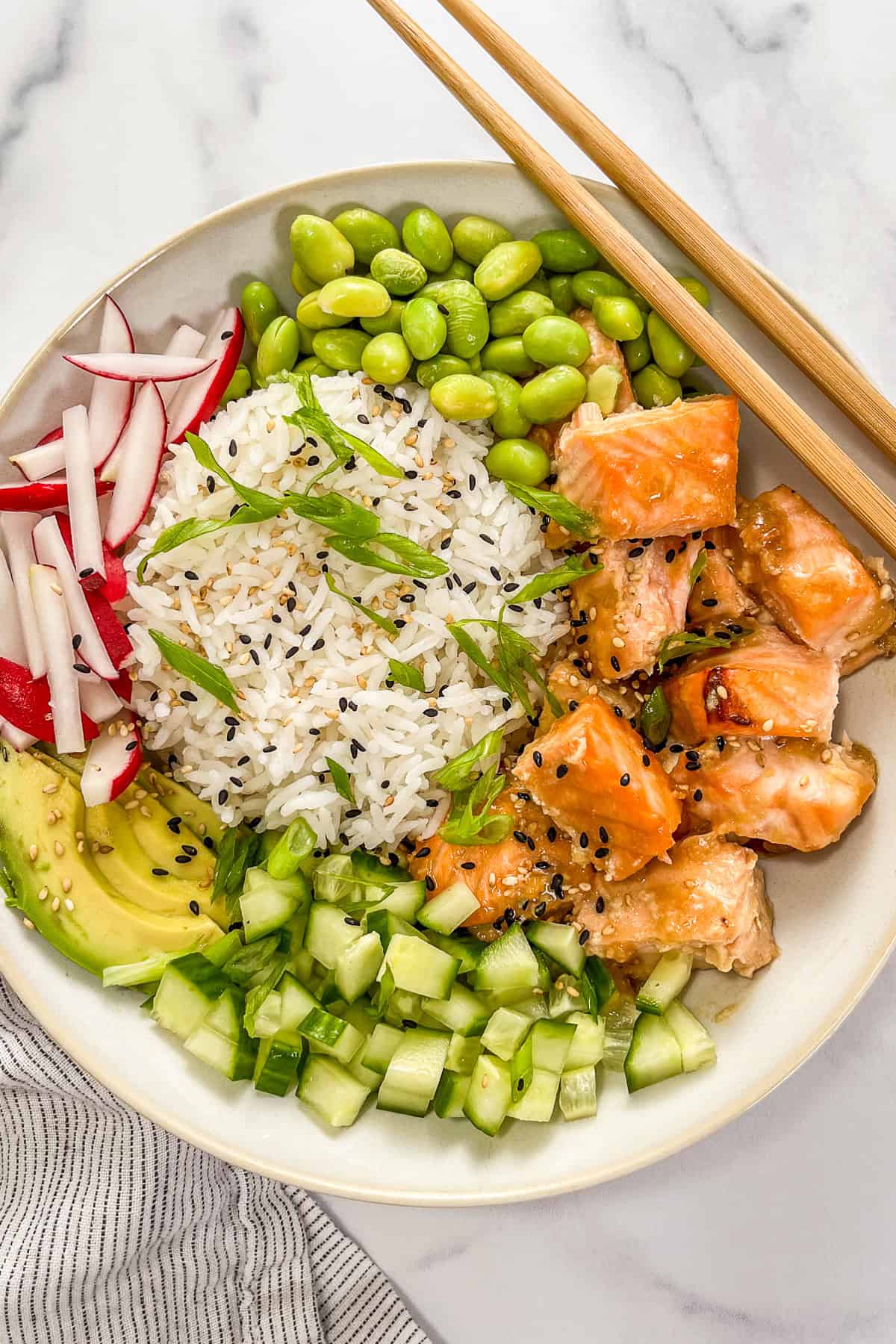Miso salmon rice bowl with edamame, sliced green onions, sliced radishes, sliced avocado, chopped cucumber, and a pair of wooden chopsticks; next to a striped towel.