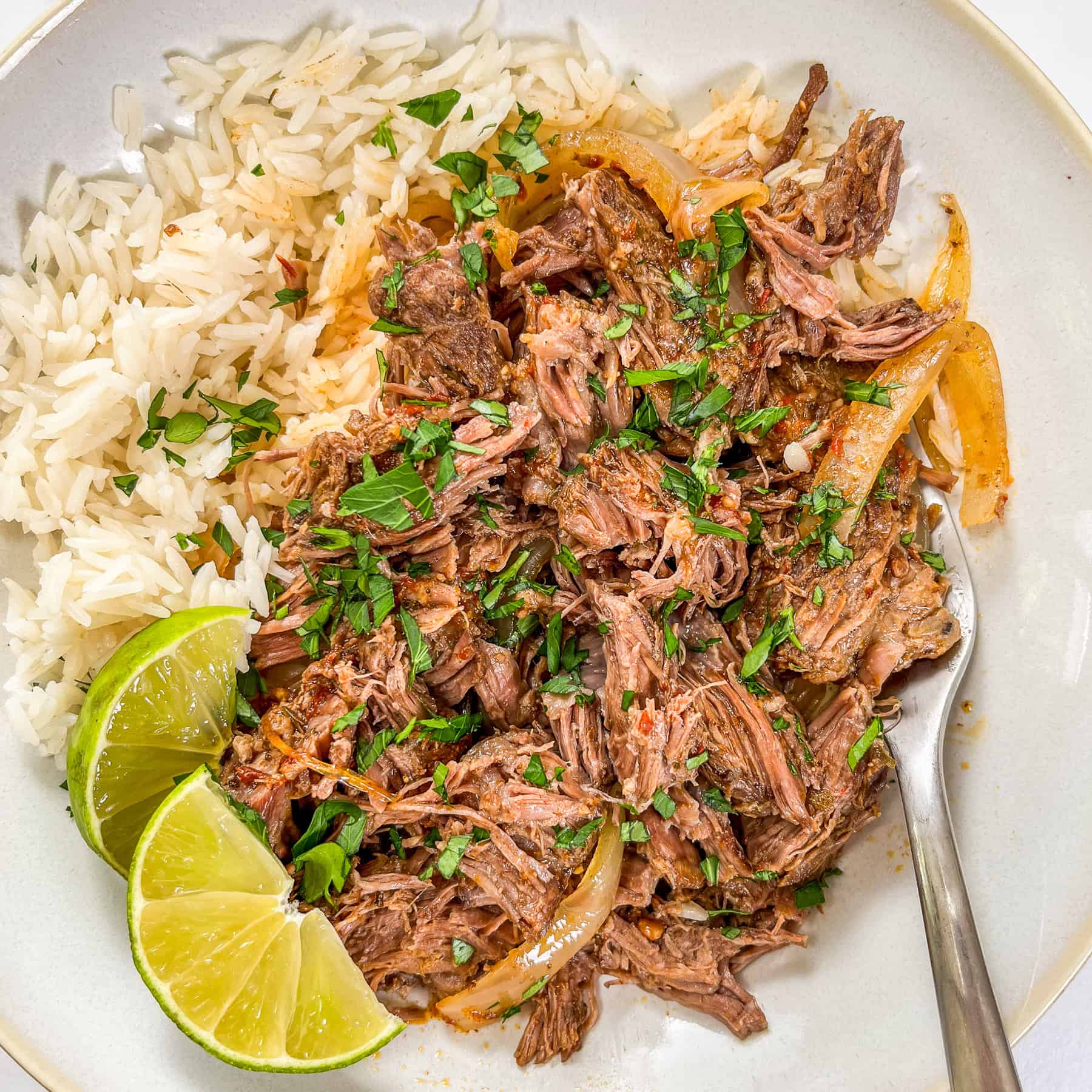 Shredded beef barbacoa with rice and lime wedges in a white bowl with a silver fork.