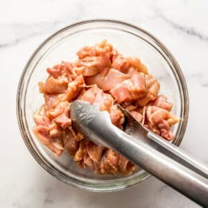Chicken chunks being tossed with corn starch in a glass bowl with silver tongs.