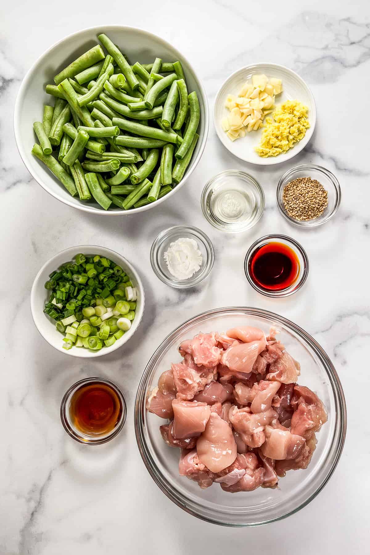 Ingredients for green bean and chicken stir fry in bowls on a marble background.