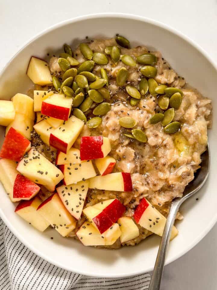 Pumpkin spiced oatmeal with apples, pepitas, chia seeds, and honey ini a white bowl with a spoon.