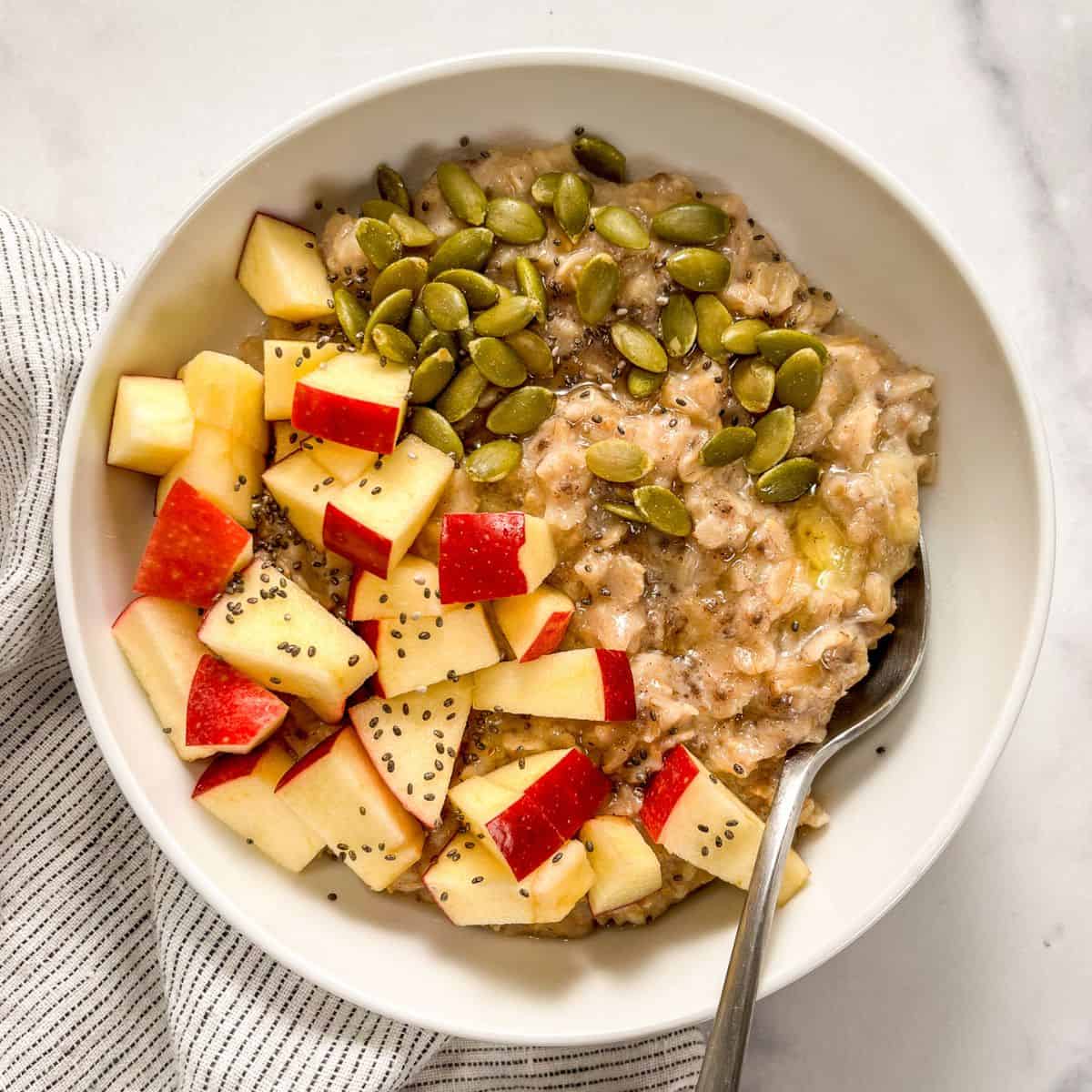 Pumpkin spiced oatmeal with apples, pepitas, chia seeds, and honey ini a white bowl with a spoon.