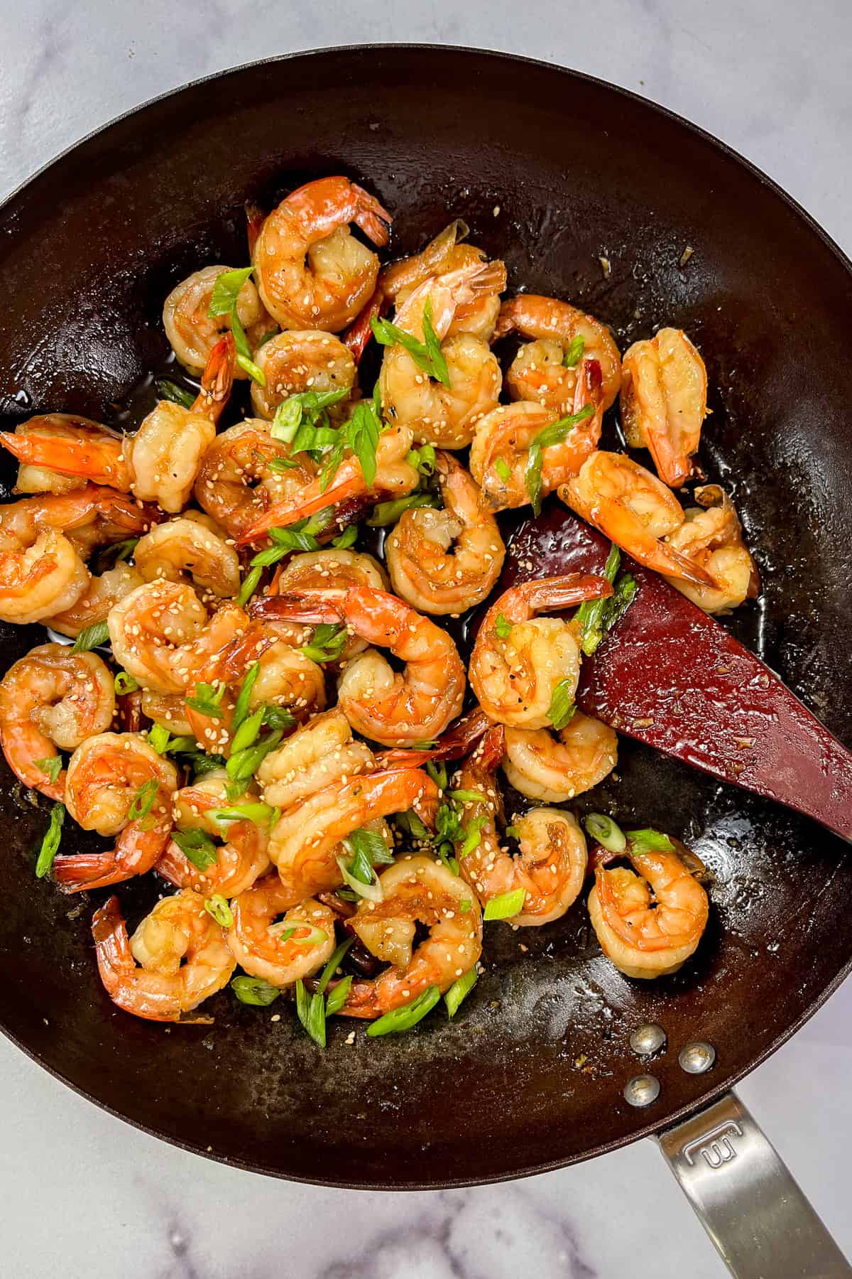 Honey garlic shrimp topped with sesame seeds and sliced green onions in a large pan with a wooden spoon.