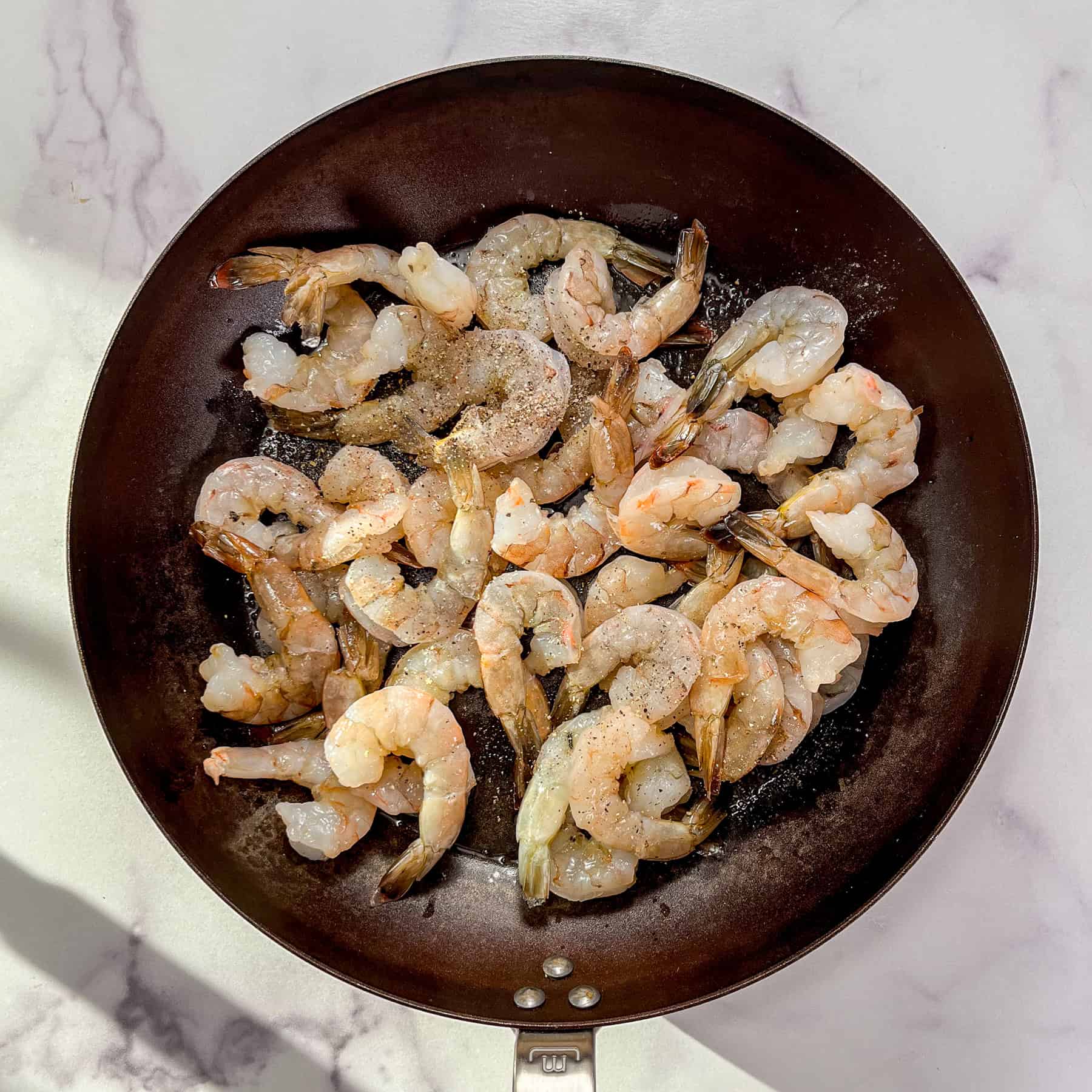 Raw shrimp topped with salt and pepper in a frying pan.