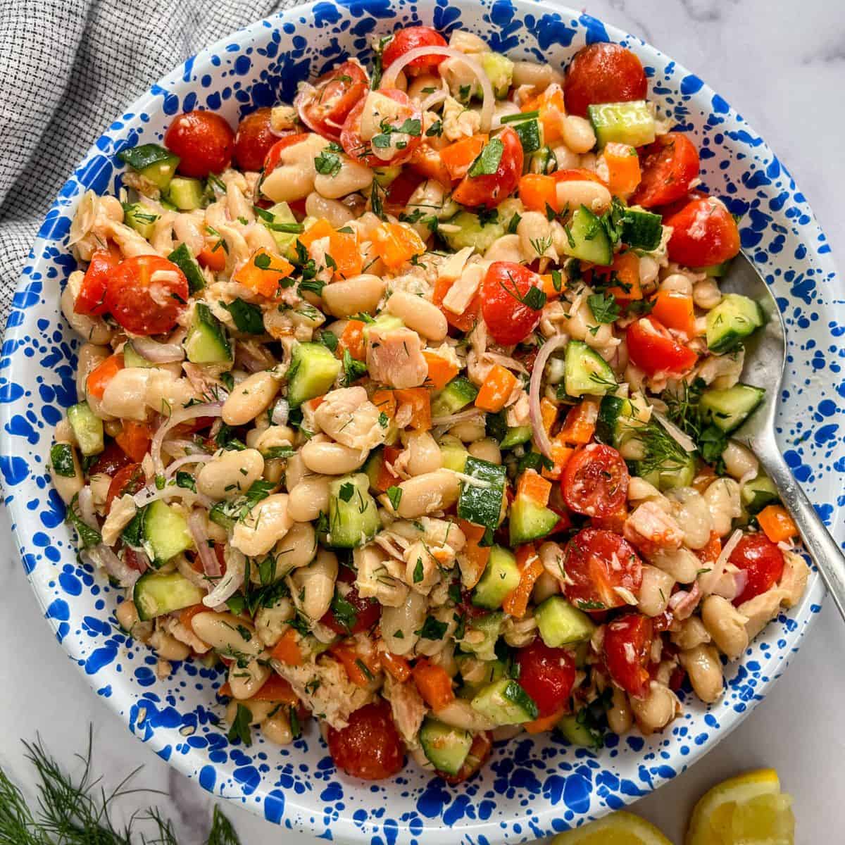 Tuna white bean salad with vegetables and herbs in a blue and white speckled bowl with a silver spoon.
