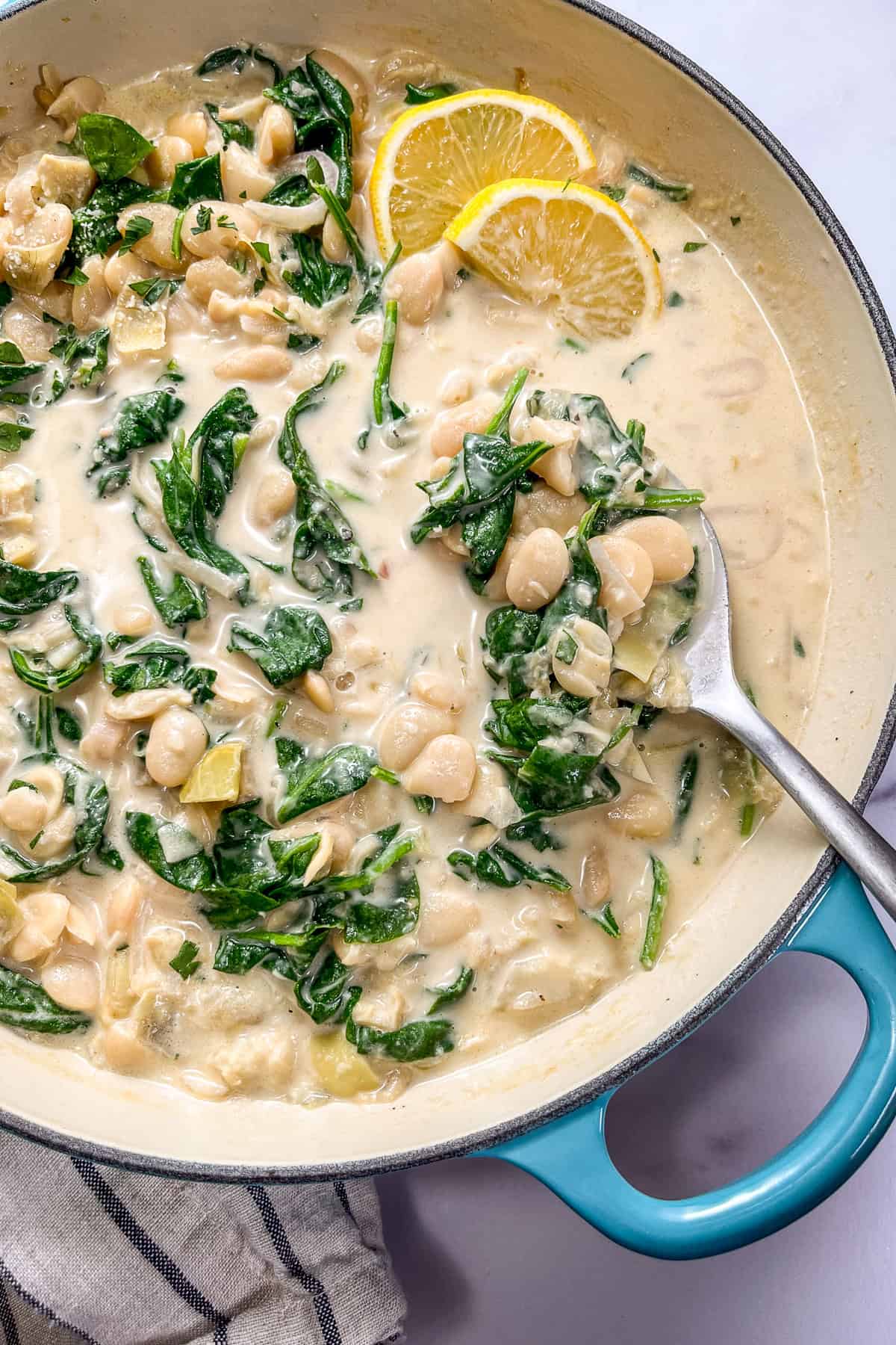 Spinach artichoke butter beans in a creamy sauce in a pan with a silver spoon and sliced lemons.