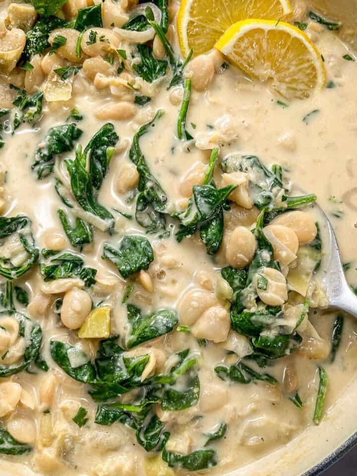 Spinach artichoke butter beans in a pan with a spoon and sliced lemons.