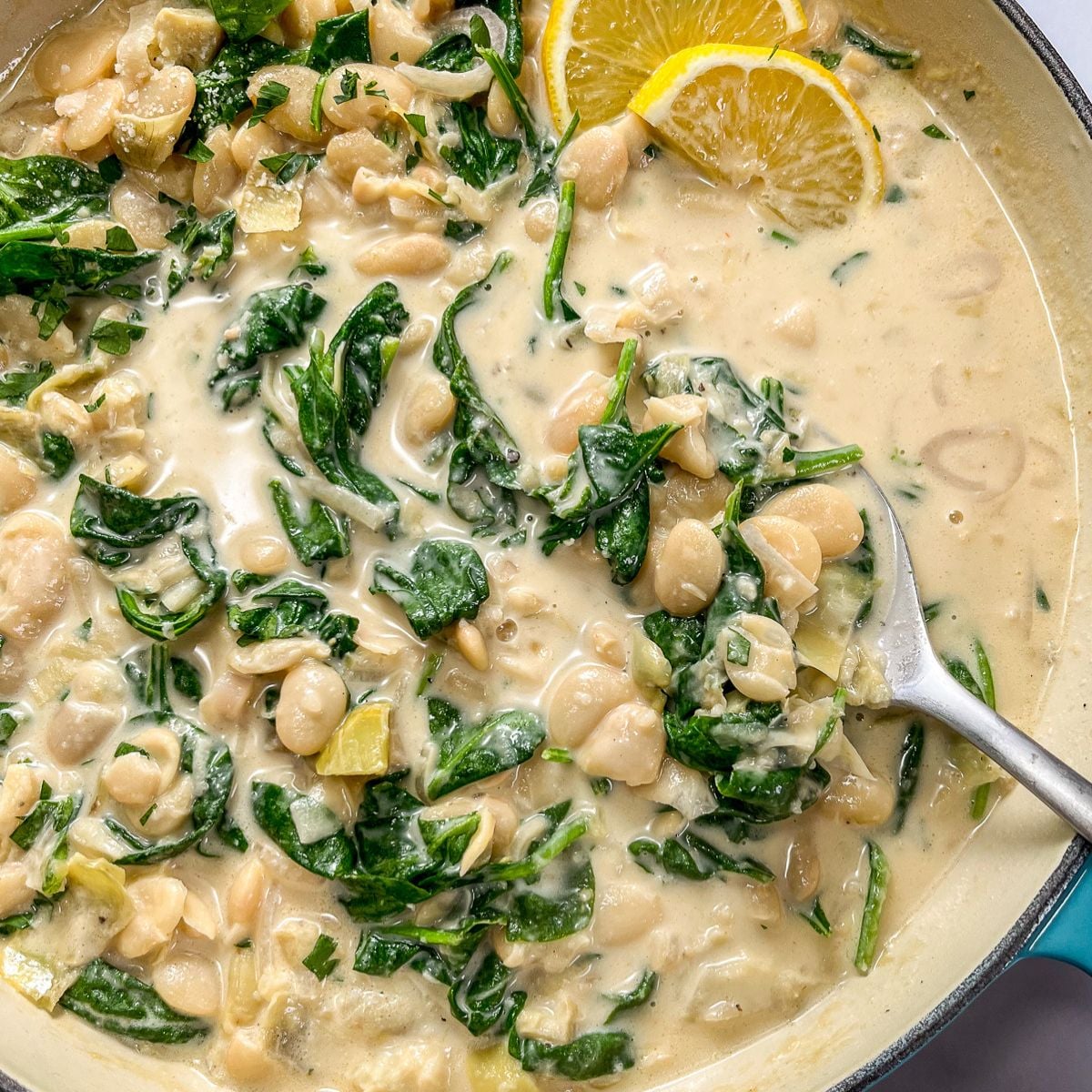 Spinach artichoke butter beans in a pan with a spoon and sliced lemons.