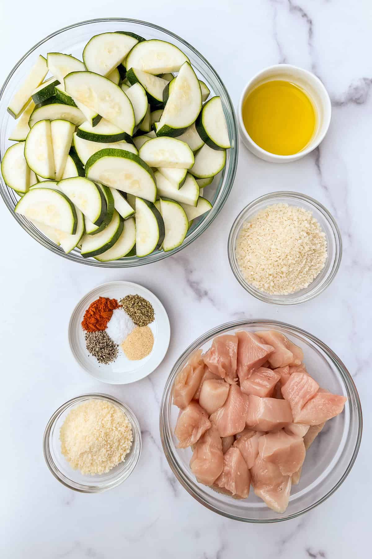 Zucchini, olive oil, parmesan, panko, spices, and chicken pieces in bowls.