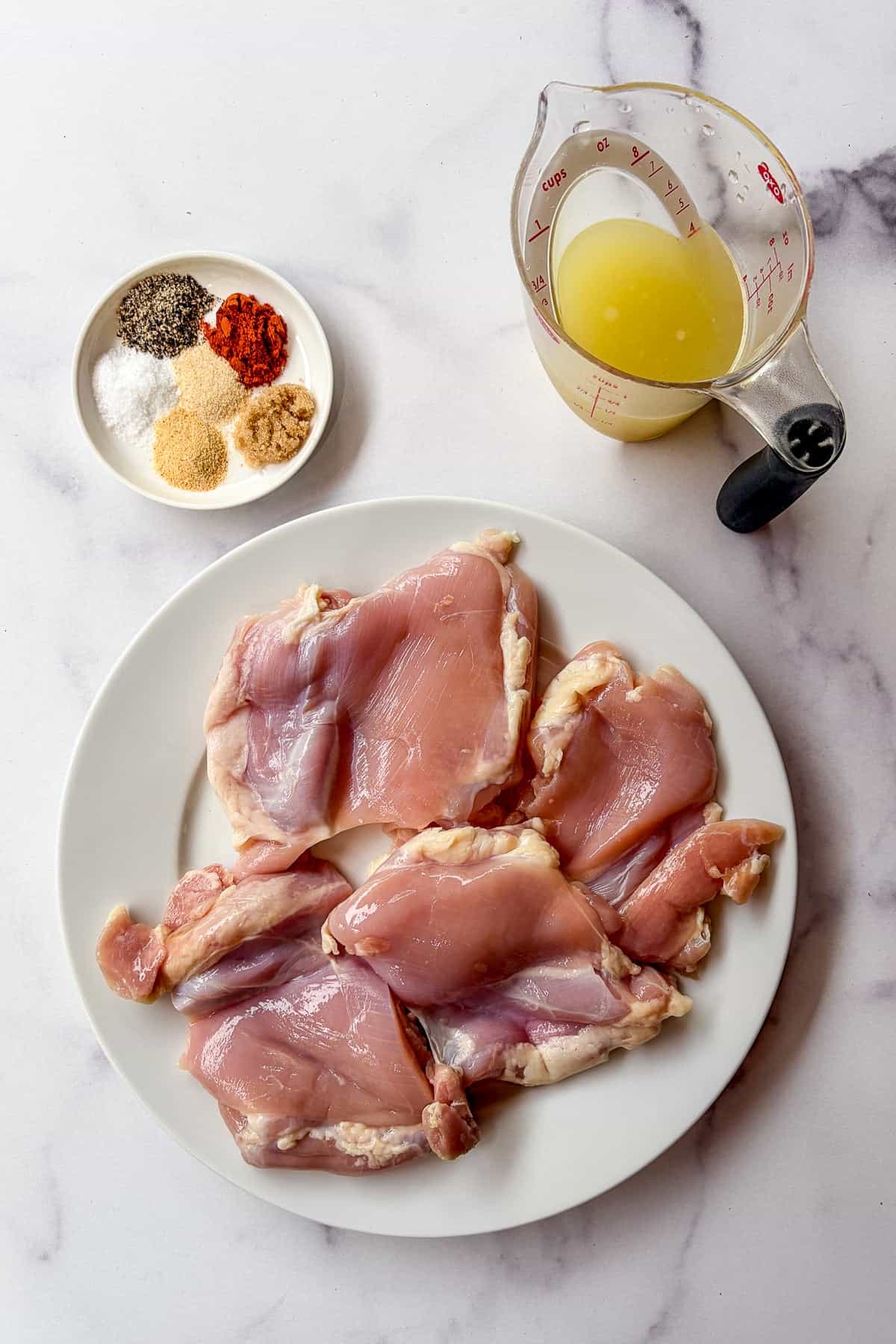 Raw chicken thighs on a white plate next to a measuring cup of chicken stock and a small plate of spices.